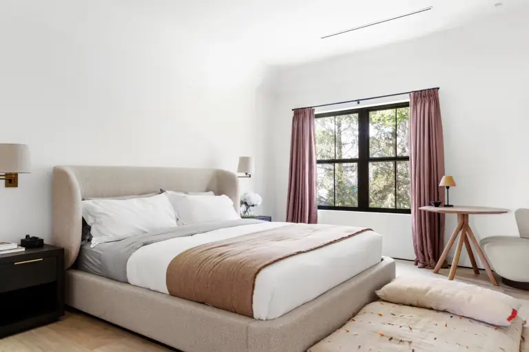 Bedroom with an upholstered platform bed, brass sconces and mauve drapes over a black-framed window at the Farmstead