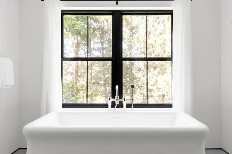 Freestanding tub with chrome fixtures before a black-framed window overlooking the pines at the Farmstead