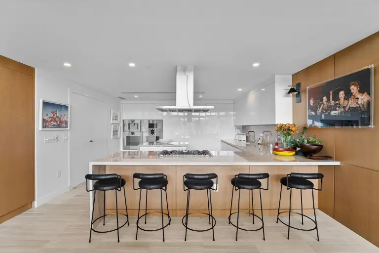 Kitchen with a marble island on an oak base, black bar stools and glossy white cabinetry at Starr DeVos Condo