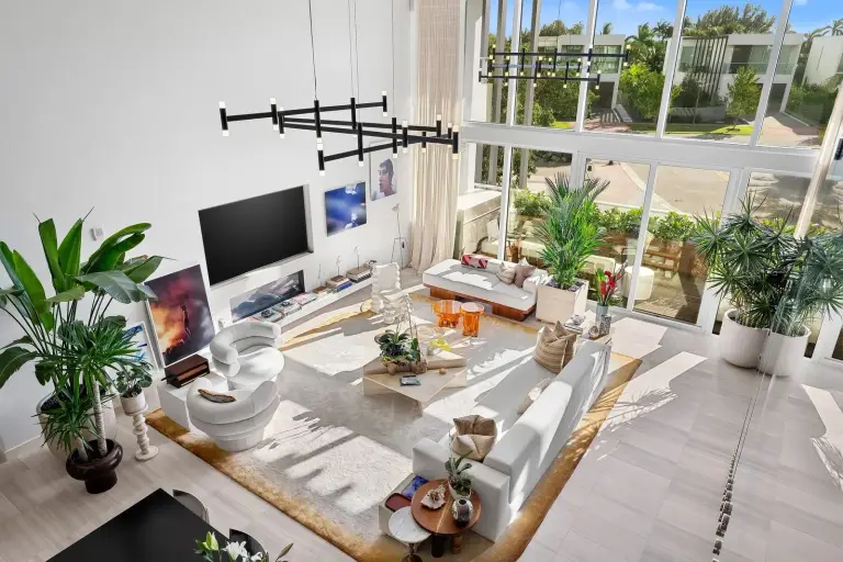 Double-height living room seen from the mezzanine with white sofas, palms and a sculptural chandelier at Starr DeVos Condo